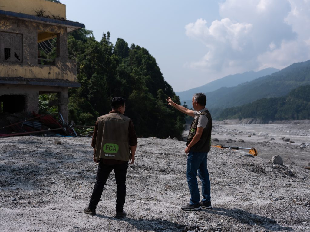 Two people in FCA branded vests stand in front of a ruined building in a devastated and mud-filled landscape. In the background we see mountains.